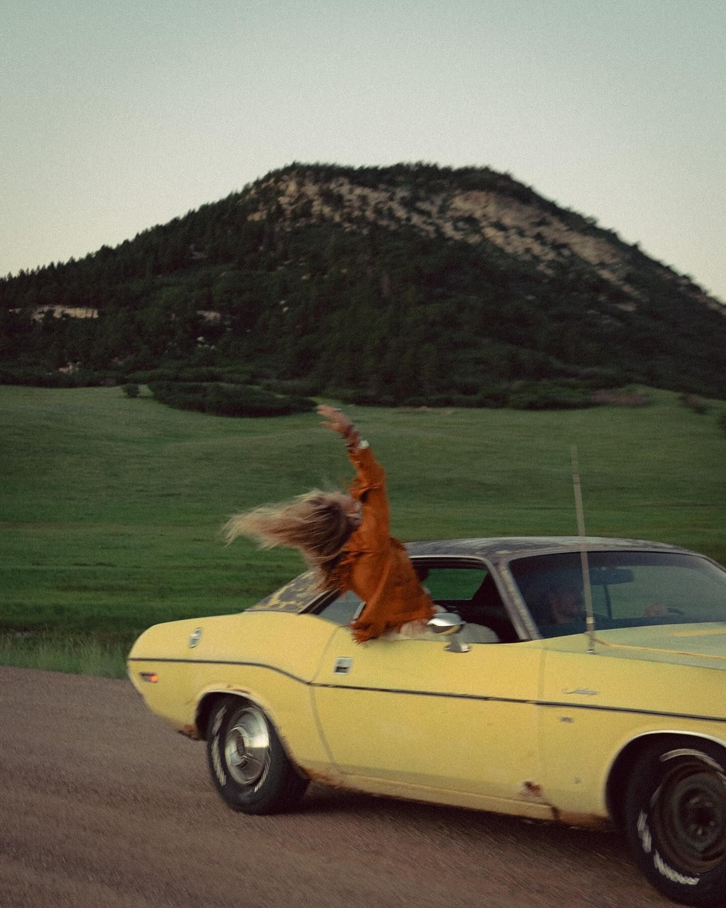 A woman joyfully reaching from the window of a moving vintage car